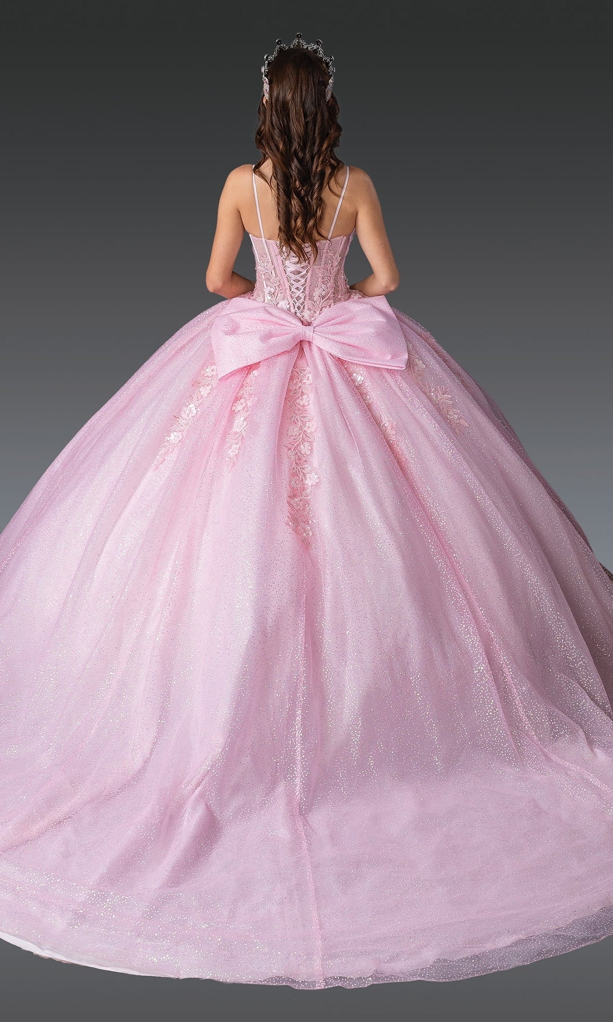 A person stands facing away against a plain dark background, wearing the Dancing Queen 1958 long quinceañera dress in pink, featuring a large bow on the back.